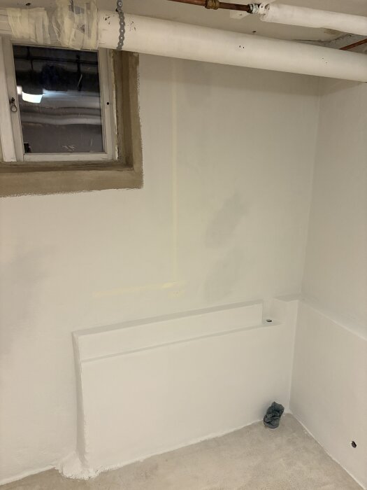 Nymålad vit vägg i en tvättstuga med ett fönster och rör synliga, en förlängd betongklack och TM-avlopp i hörnet.