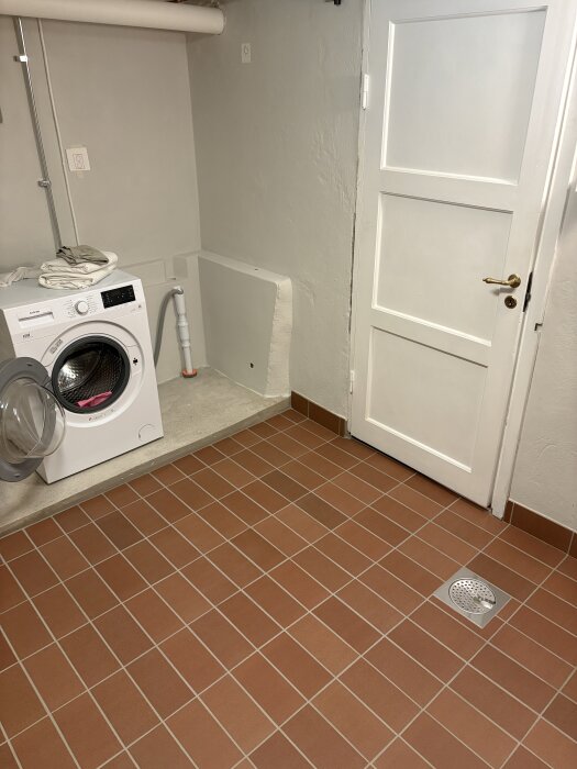 Tvättstuga med tvättmaskin, klinkergolv i rött, vit dörr och synligt avlopp i hörnet. Betongklack förlängd, väggar målade i ljus färg.