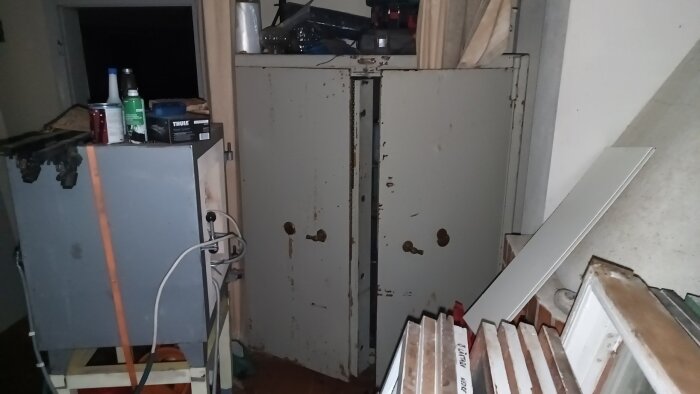 En grå keramikugn med anslutna kablar och ett stort, slitet kassaskåp i bakgrunden, omgiven av byggmaterial och verktyg.