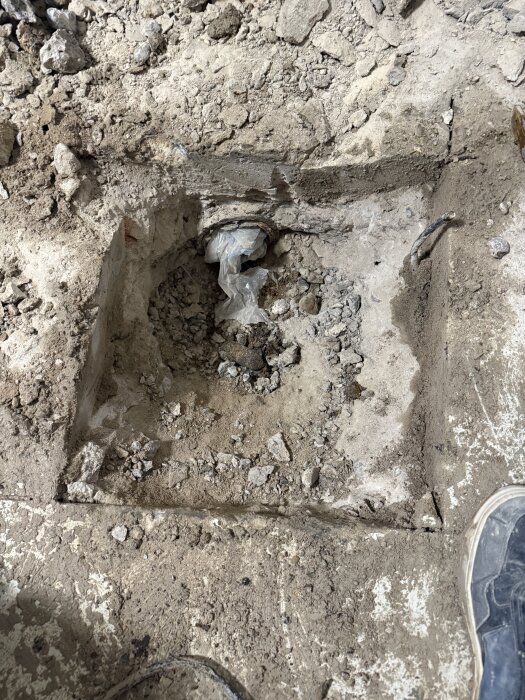 Grop i golvet där gamla brunnar har avlägsnats, omgiven av grus och jord. En sko syns i hörnet av bilden.
