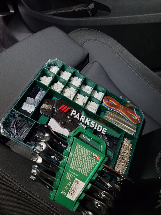 Crimptång och en uppsättning spärrnycklar i ett bilsäte, omgiven av diverse små komponentsatser i en grön förvaringsbox.