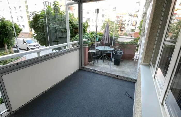 Inglasad balkong med glasräcke, möblerad med bord och stolar, omgiven av krukväxter, i stadsbebyggelse.