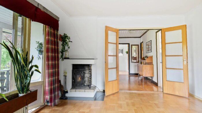 Renoverat vardagsrum med öppen spis, trägolv och glasdörrar, växter vid fönstret och gardiner.