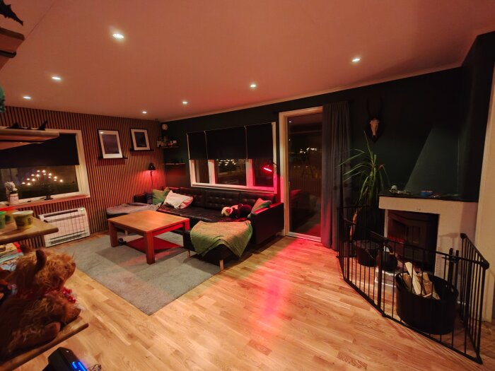 Renoverat vardagsrum med trägolv, svart soffa, träbord, gröna väggar, inbyggda högtalare, och en eldstad omgiven av vedkorgar.