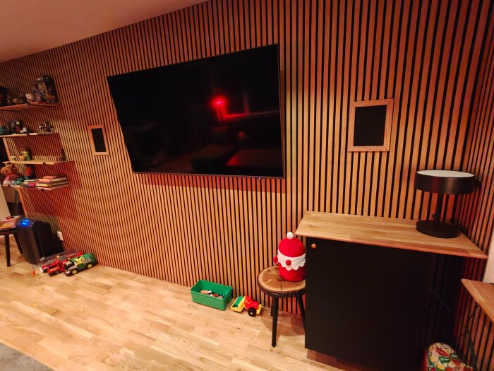 TV monterad på ribbvägg i nyrenoverat vardagsrum med infällda högtalare. Hyllor med leksaker och en lampa på ett skåp syns också.