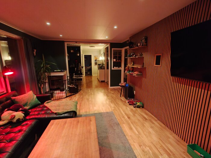 Renoverat vardagsrum med infälld TV och hyllor, nya träväggar och golv, samt modern belysning. Lekfull inredning med leksaker i hörnet.