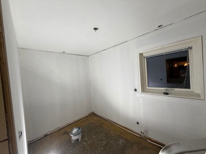 Rum med grundmålade vita väggar, ett fönster utan gardiner och en burk målarfärg på golvet.