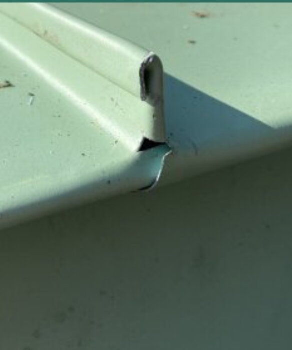 Närbild av en skadad grön plåt där kanten är böjd och skadad, med synliga repor och märken.