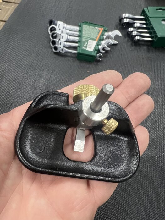 Hand håller en liten hyvel med metall- och gulddetaljer, i bakgrunden syns en nyckelsats på en svart matta.