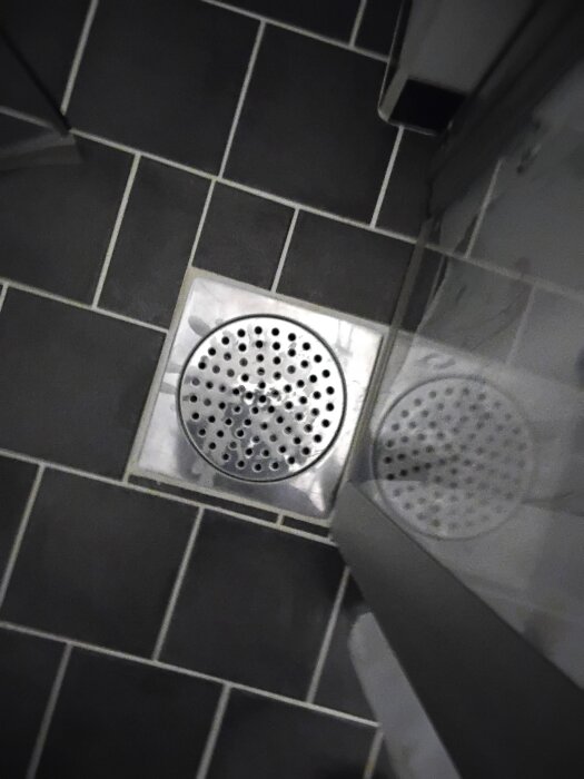 Golvbrunn i dusch med mörk klinker och metallgallerlock.