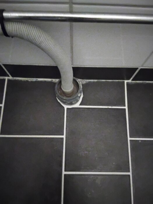 Grå plaströr kopplat till avlopp mot svartvit kaklad vägg och golv.