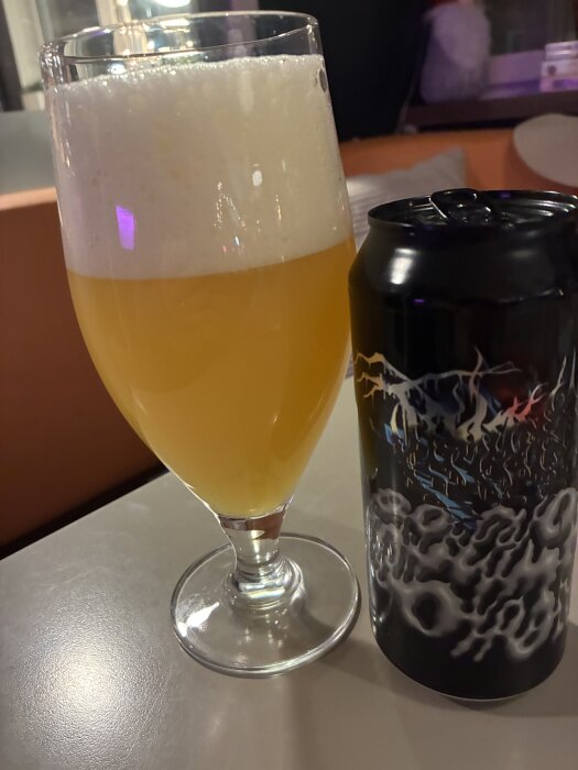 Ölglas med skummande bärnstensfärgad dryck och en dekorerad ölburk på ett bord.