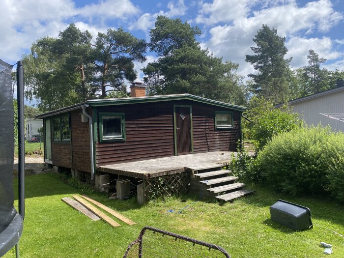 Trästuga med övergiven altan, omgiven av träd och buskar. Synliga plankor och en liggande svart behållare på gräsmattan.