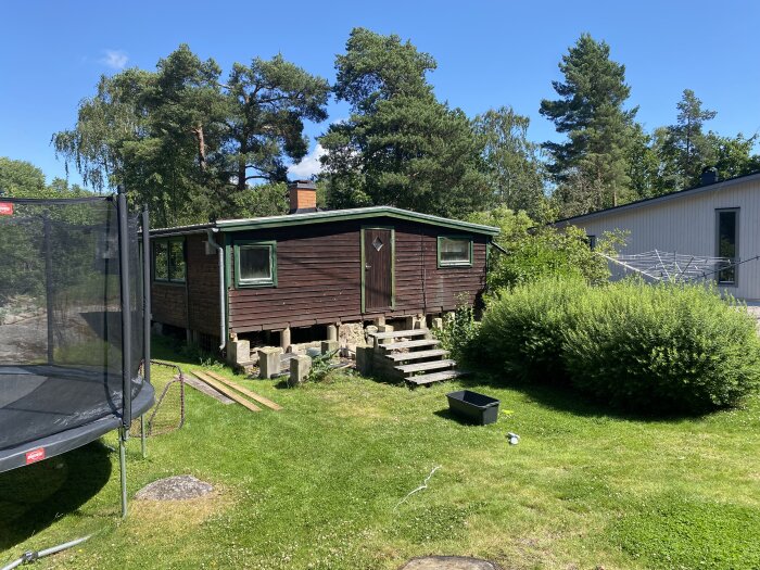 En trästuga utan altan, omgiven av gräs och buskar, bredvid en studsmatta.