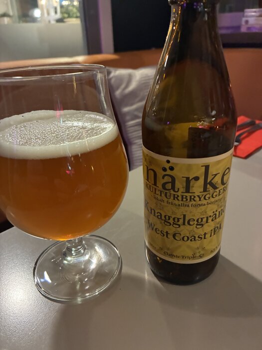 Ett glas med skummande West Coast IPA från en flaska märkt "Närke Kulturbryggeri" på ett bord i en avslappnad miljö.
