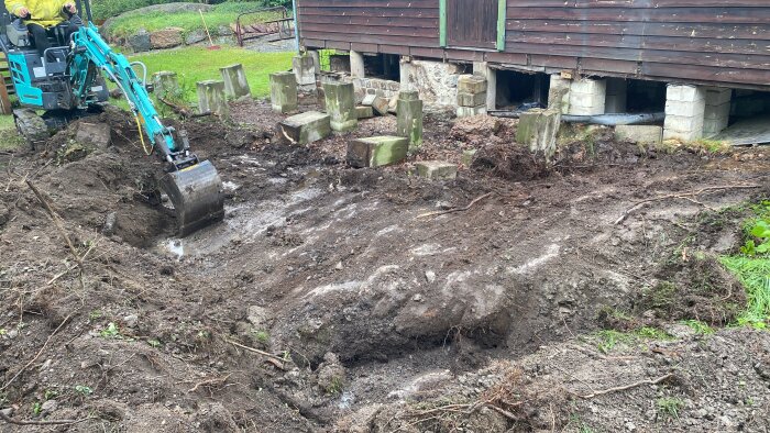 Grävmaskin gräver vid ett trähus på plintar, gör förberedelser för markarbete, med jord och gräs runtom.