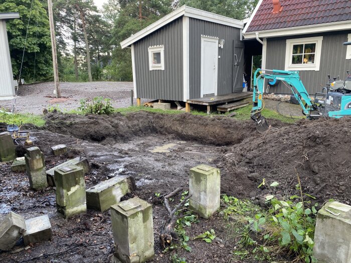 En minigrävare gräver ut en grund intill ett grått hus med vita detaljer. Betongblock ligger utspridda runt den uppgrävda jorden.