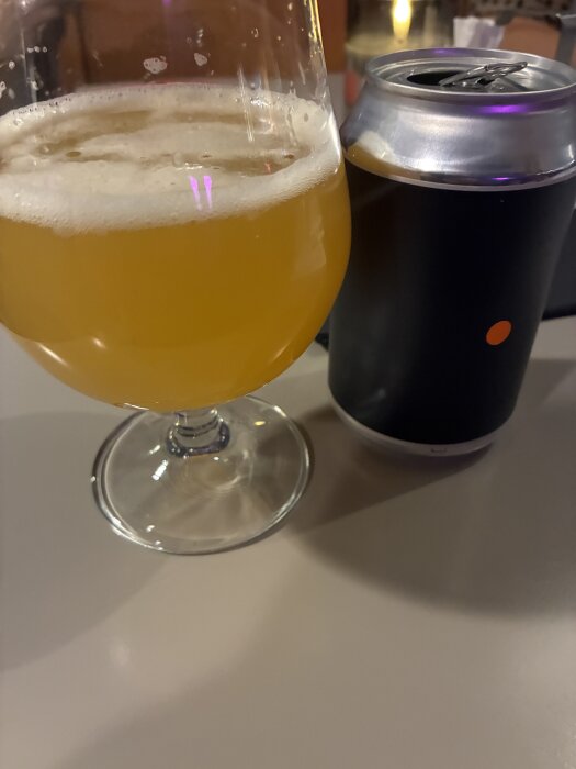 Ett glas halvt fyllt med ljus öl står bredvid en mörk aluminiumburk på ett bord.