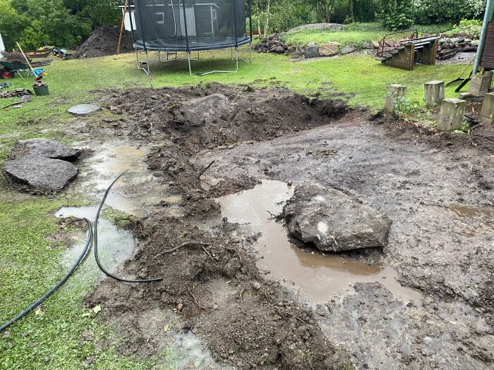 Uppgrävd trädgård med lera och stenar, vattenpölar och en trampolin i bakgrunden.