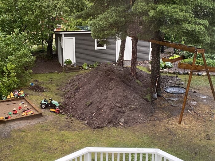 Lekplats med sandlåda och leksaksfordon, jordhög under träd vid gunga, grönska och förråd i bakgrunden.