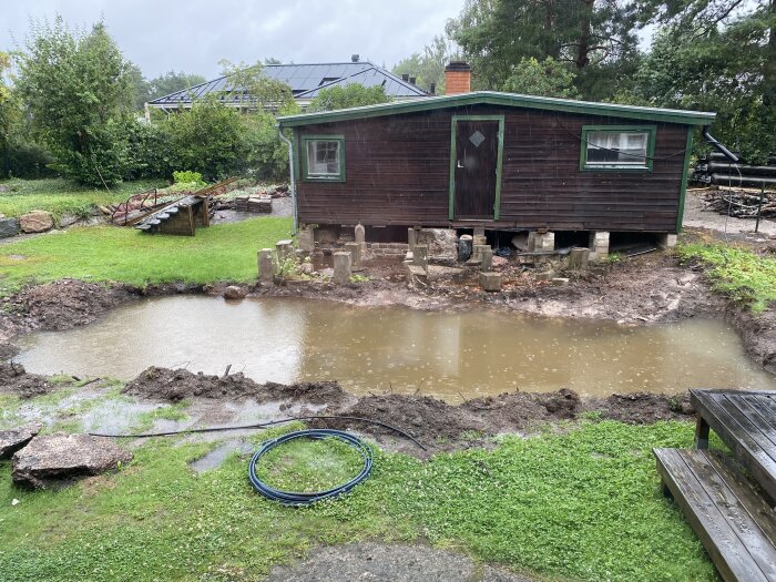 Stor vattenfylld grop framför en brun stuga, omgiven av grönska, efter kraftigt regn. En blå slang ligger på den blöta gräsmattan.