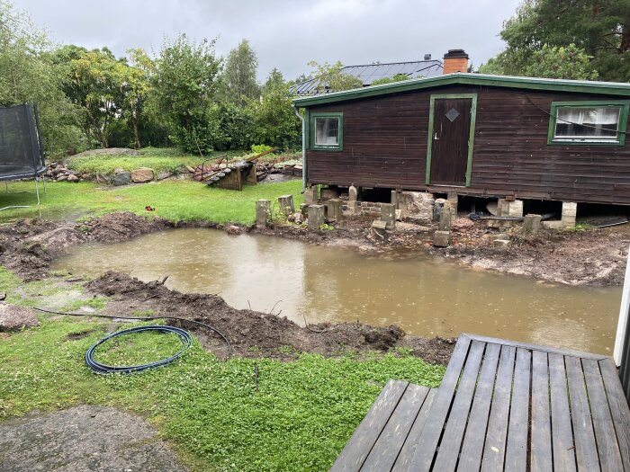 En vattenfylld grop framför ett upphöjt trähus och grön gräsmatta, med trampolin i bakgrunden.
