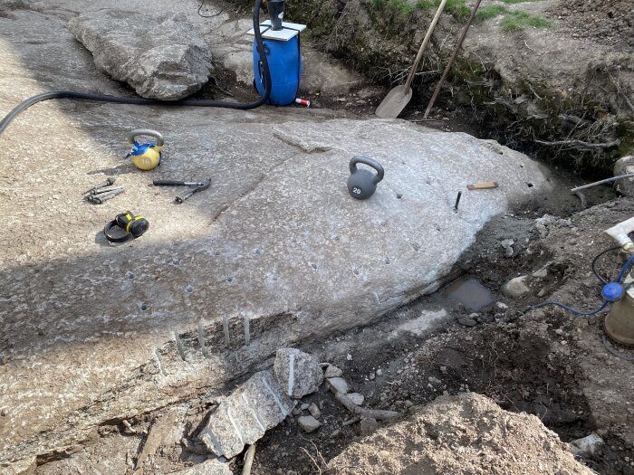 Bergyta med borrhål, vikter, verktyg och spräckningsutrustning för snigeldynamitprocess, omgiven av jord och gräsmatta.