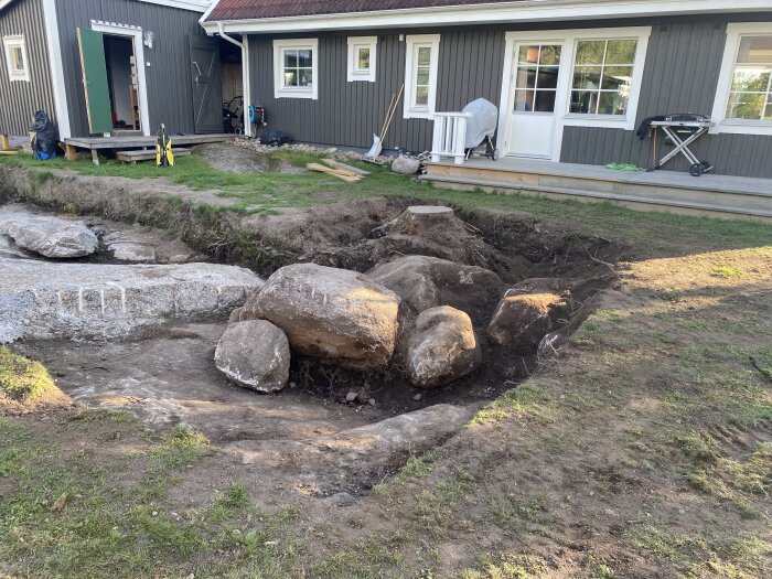 Grävd yta vid hus med stora stenar och stubb, tydlig nivåskillnad i marken.