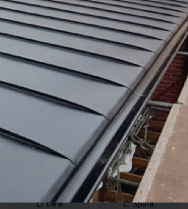 Svart plåttak med stående sömmar under installation. Ställning och tegelvägg ses i bakgrunden.