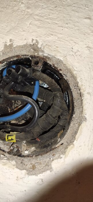 Öppnad väggdosa i källare med gamla och nya kablar, inklusive svart och blå kabel, synliga i en sliten vägg.