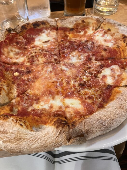 En rund pizza med tomatsås och smält ost på en tallrik, med dryckesglas i bakgrunden.