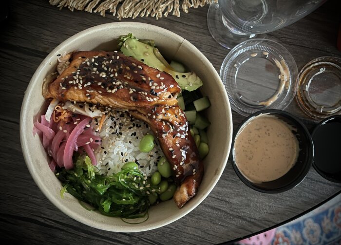 Pokébowl med teriyakilax, avokado, edamame, sjögräs, ris och picklad rödlök, serverad i en beige skål med såser vid sidan.