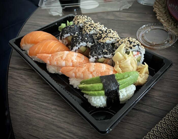Sushi-förpackning med nigiri av lax och räka samt makirullar med sesamfrön, dekorativt upplagt med avokado och ingefära på ett träbord.