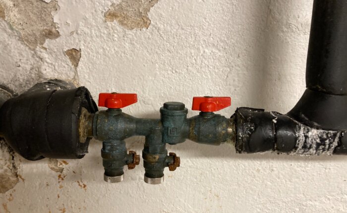 Rörinstallation med två röda ventiler och isolering, monterad på en vit, sliten vägg.