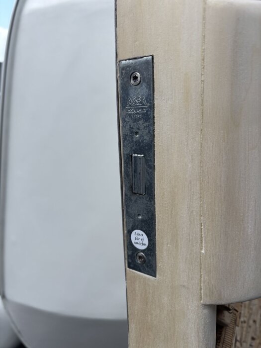 Närbild på en dörrsida med ett monterat metallås från märket ASSA. Låset har en etikett med texten "Låset får ej smörjas".