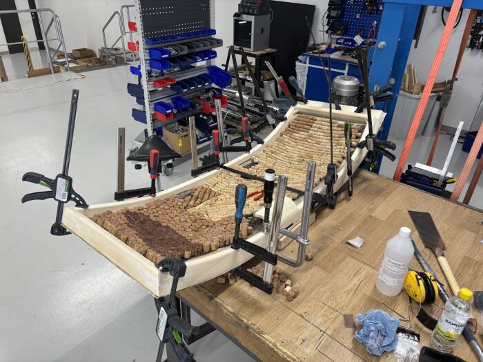 En dörrkarm under limning med korkar fastsatta och flera tvingar på plats. Verktyg och material syns på arbetsbänken i verkstaden.