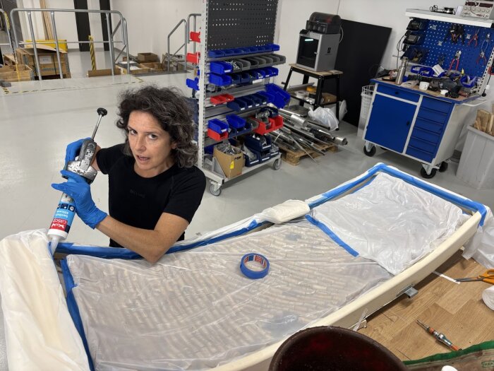 Person applicerar silikon med fogspruta på dörryta i verkstad, omgiven av verktyg och material.