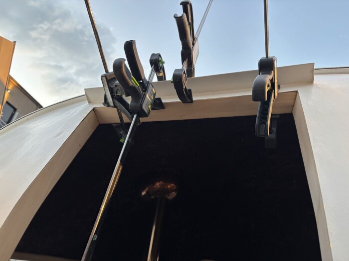 Trälister pressas på plats med tvingar vid dörröppning under limningsprocessen i byggprojekt, mot bakgrund av en klar himmel.