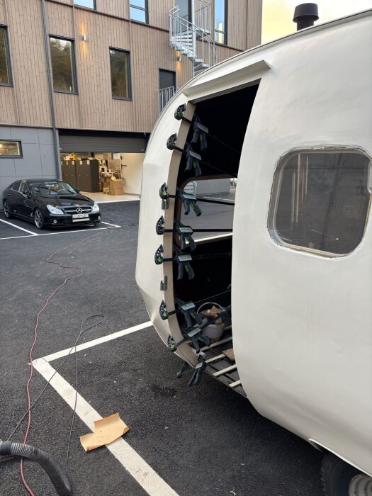 Husvagn under renovering med träkarm och flera tvingar som håller delarna på plats, står parkerad utanför en byggnad.