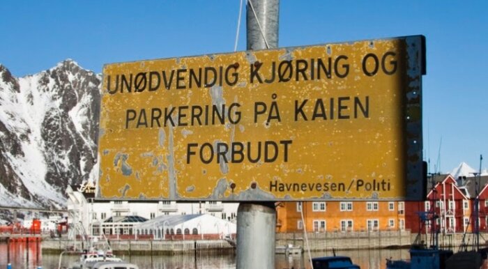 Sliten skylt vid hamn med texten "UNØDVENDIG KJØRING OG PARKERING PÅ KAIEN FORBUDT", snöiga berg och färgglada byggnader i bakgrunden.