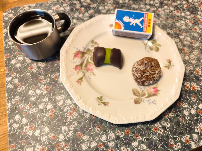 Chokladdoppad grön marsipanbakelse och kokosboll på blommig tallrik, med en kopp kaffe och Solstickan tändstickor bredvid.