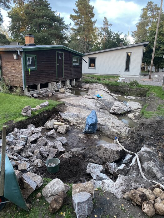 Stenar och jord runt ett mörkt hus på plintar, med trasiga stenar och verktyg kvar efter borrning, grön trädgård och andra hus i bakgrunden.