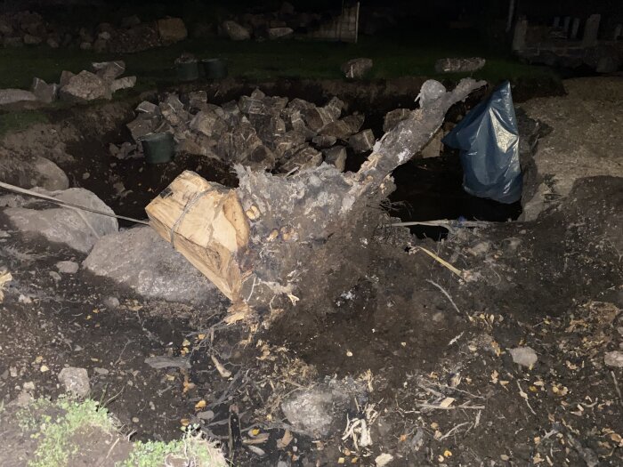 Stubbe med delvis exponerade rötter och jord. Rester av grävning och klyvning syns. Runt omkring ligger stenar och blött jord.