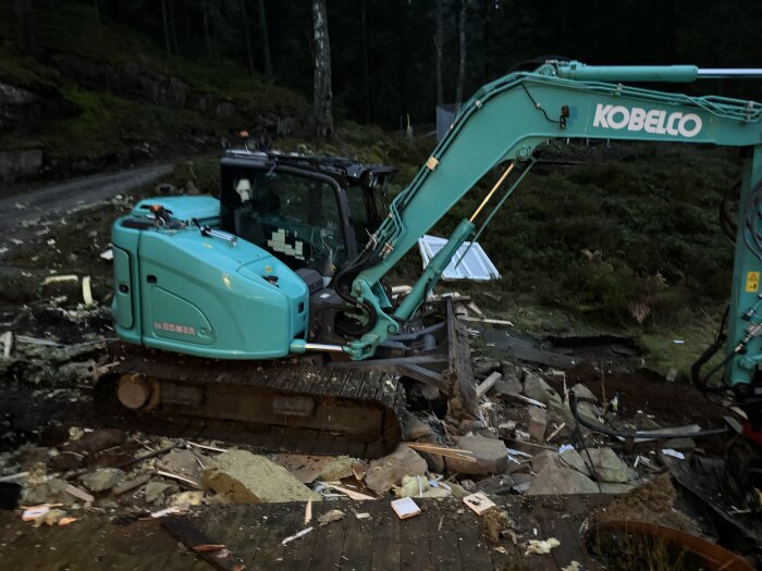Grävmaskin i arbete med markarbeten på en byggarbetsplats, omgiven av nedrivna material och sten.