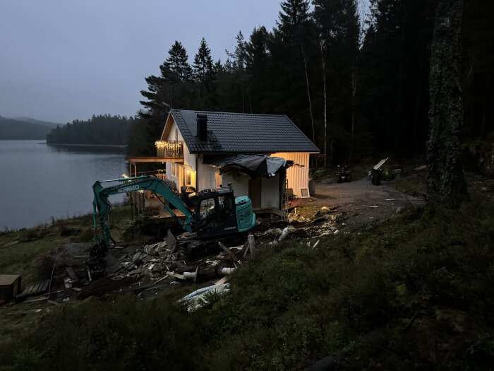 Grävmaskin framför en stuga vid en sjö i skogen, inramad av kvällsljus. Byggarbetsplats med rivningsmaterial synligt på marken.