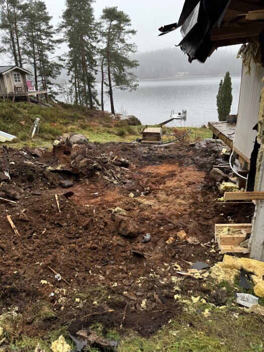 Byggarbetsplats nära sjö, markområde utgrävd med synliga stenar och trästrukturer, träd i bakgrunden och brygga vid vattnet.