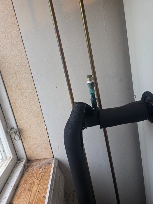 En svartisolerad rörböj med ventil bredvid två kopparrör mot en vit vägg nära ett dammigt fönster.
