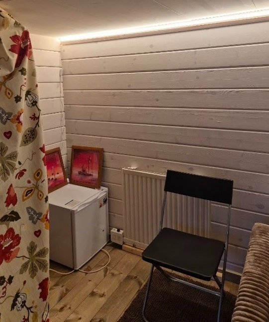 Ett rum med en vit trävägg, en stol, en fotoram, en liten vit kyl, gardin med mönster och belysning längs taklisten.