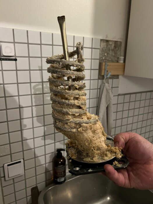 En elpatron från en varmvattenberedare täckt med kraftig kalkavlagring, hålls över en diskho med kakelvägg i bakgrunden.
