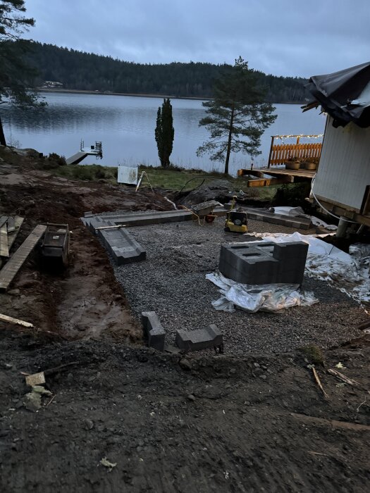 Byggarbetsplats nära sjö, med lecablock och grus. Bakgrunden visar en brygga och skogskant vid vattnet.